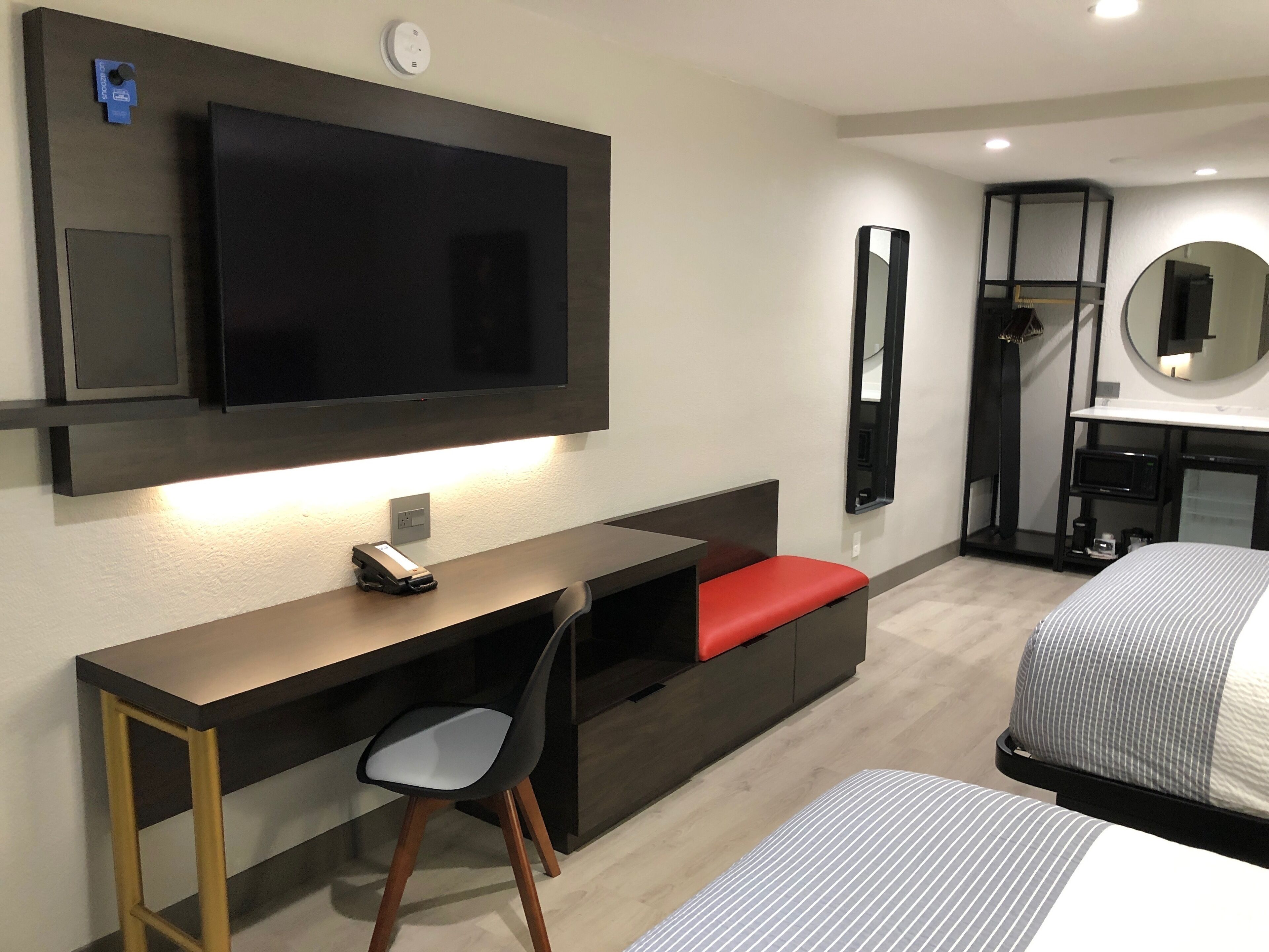 Habitación de hotel con televisión de pantalla plana montada en la pared, escritorio con silla, cama con ropa de cama a rayas, mesita de noche y espejo.