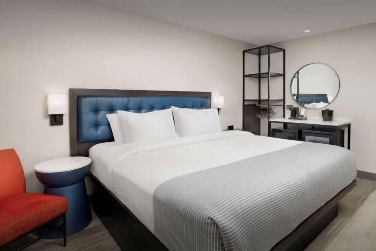 A hotel room with a bed, a red chair, a round table, a mirror, and a wall-mounted shelf.