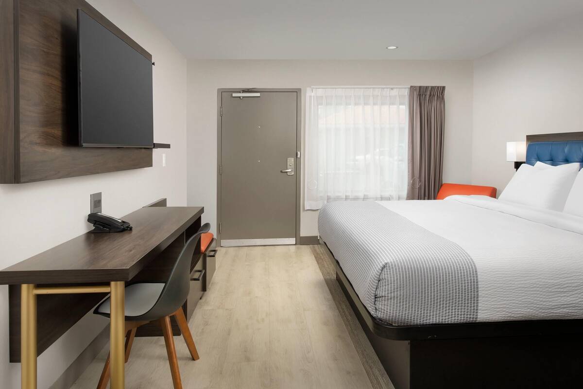 A hotel room with a bed, a desk with a telephone, a television mounted on the wall, and a door leading to another room.
