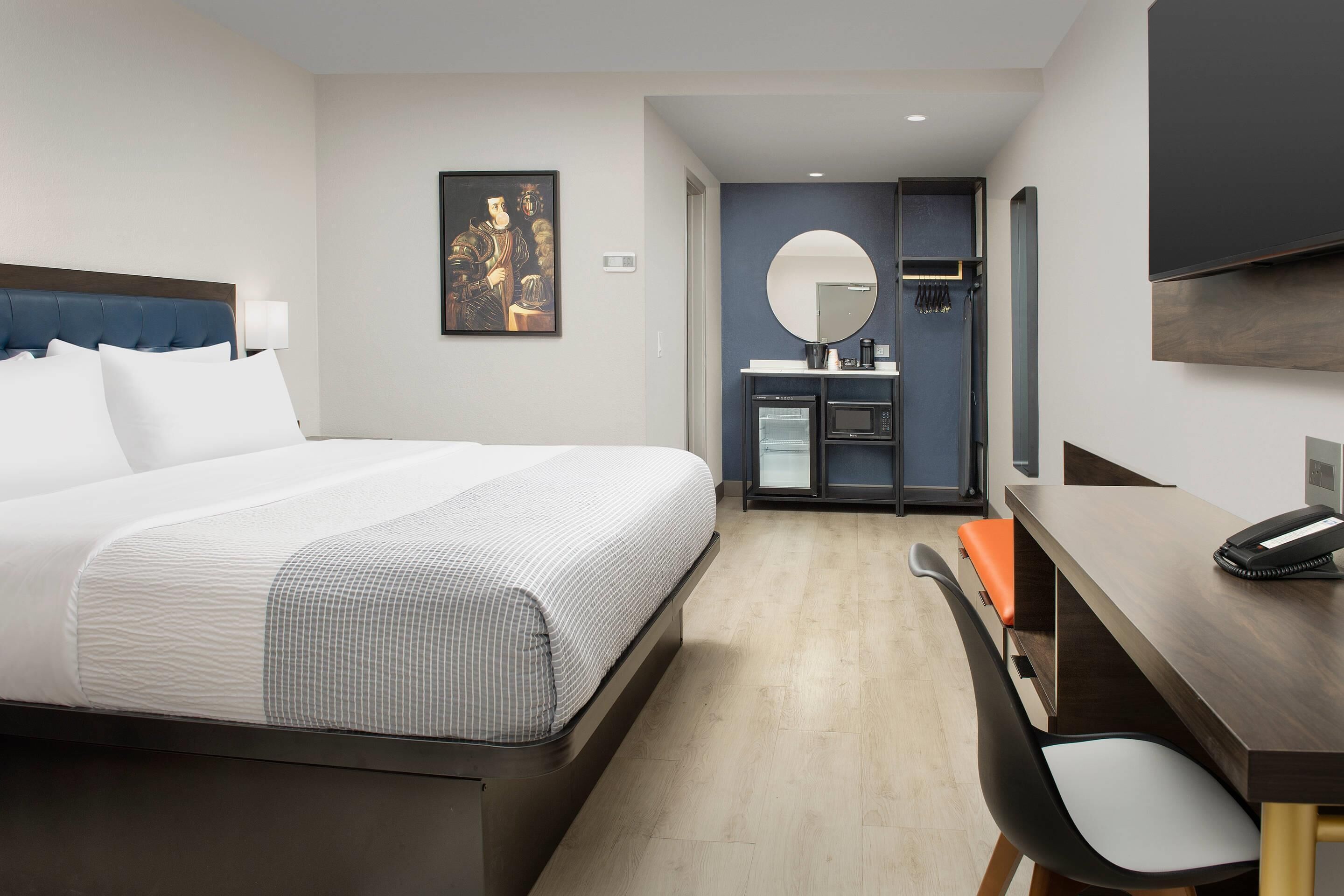 A hotel room with a bed, a desk, a chair, a mirror, and a kitchenette.