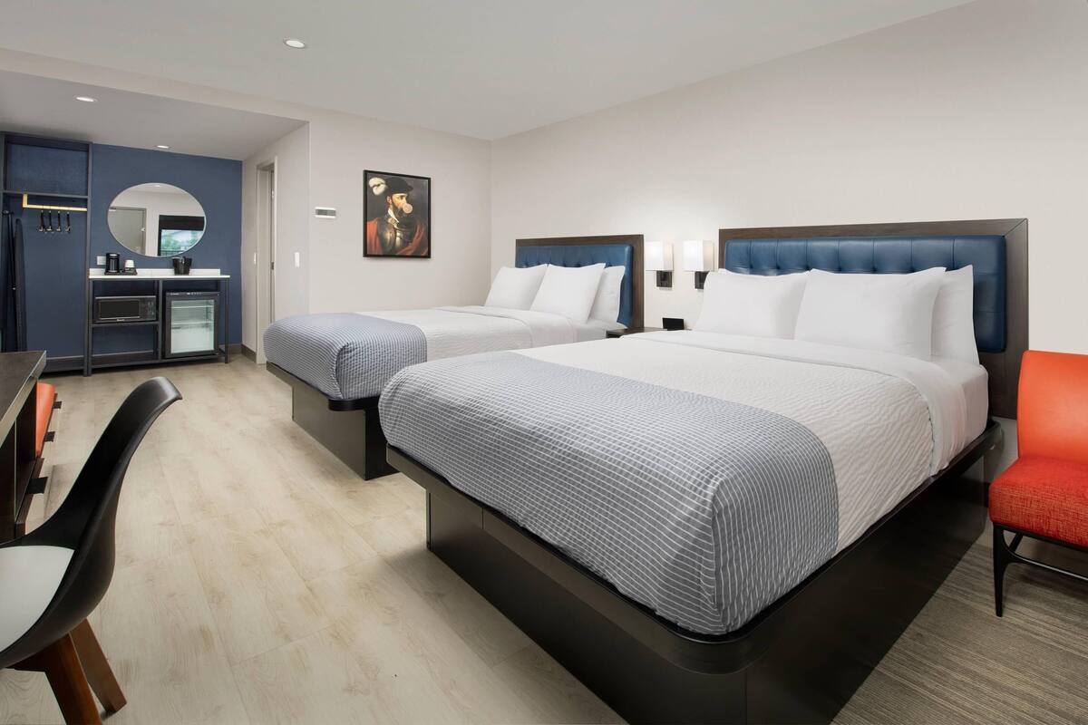 A hotel room with two beds, a desk, a chair, a microwave, and a small kitchenette.