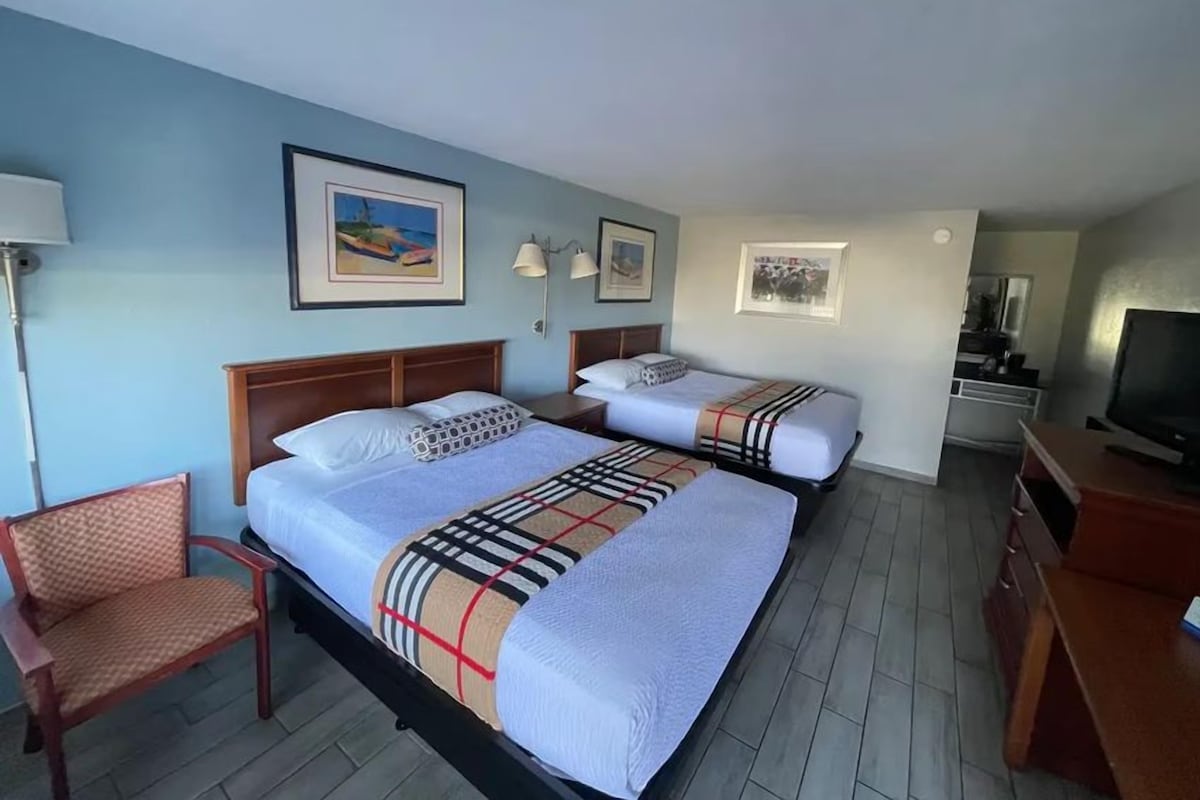 A hotel room with two beds, a chair, a desk, a TV, and artwork on the walls.