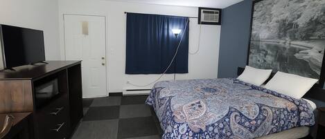 Een slaapkamer met een bed, een bureau, een televisie, een raam met gordijnen en een schilderij aan de muur.