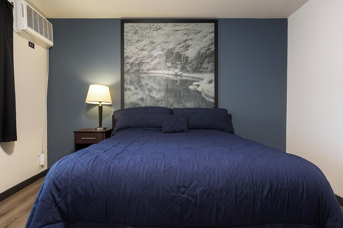 Un dormitorio con cama, mesita de noche, lámpara y un cuadro en la pared.