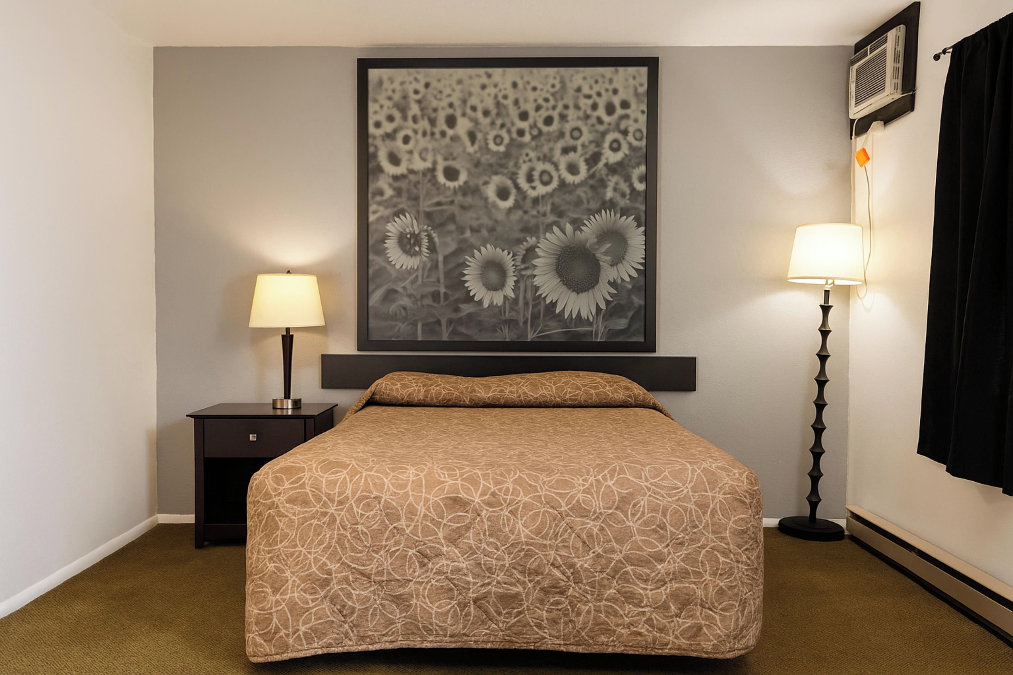 A hotel room with a bed, a nightstand, a lamp, a framed picture of sunflowers, and a window with curtains.