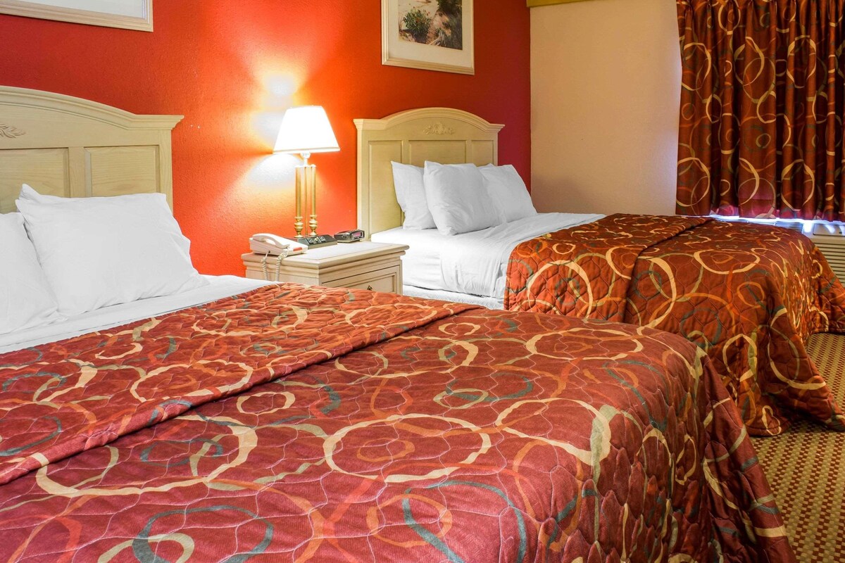 A hotel room with two beds, a nightstand with a lamp, and a window with curtains.