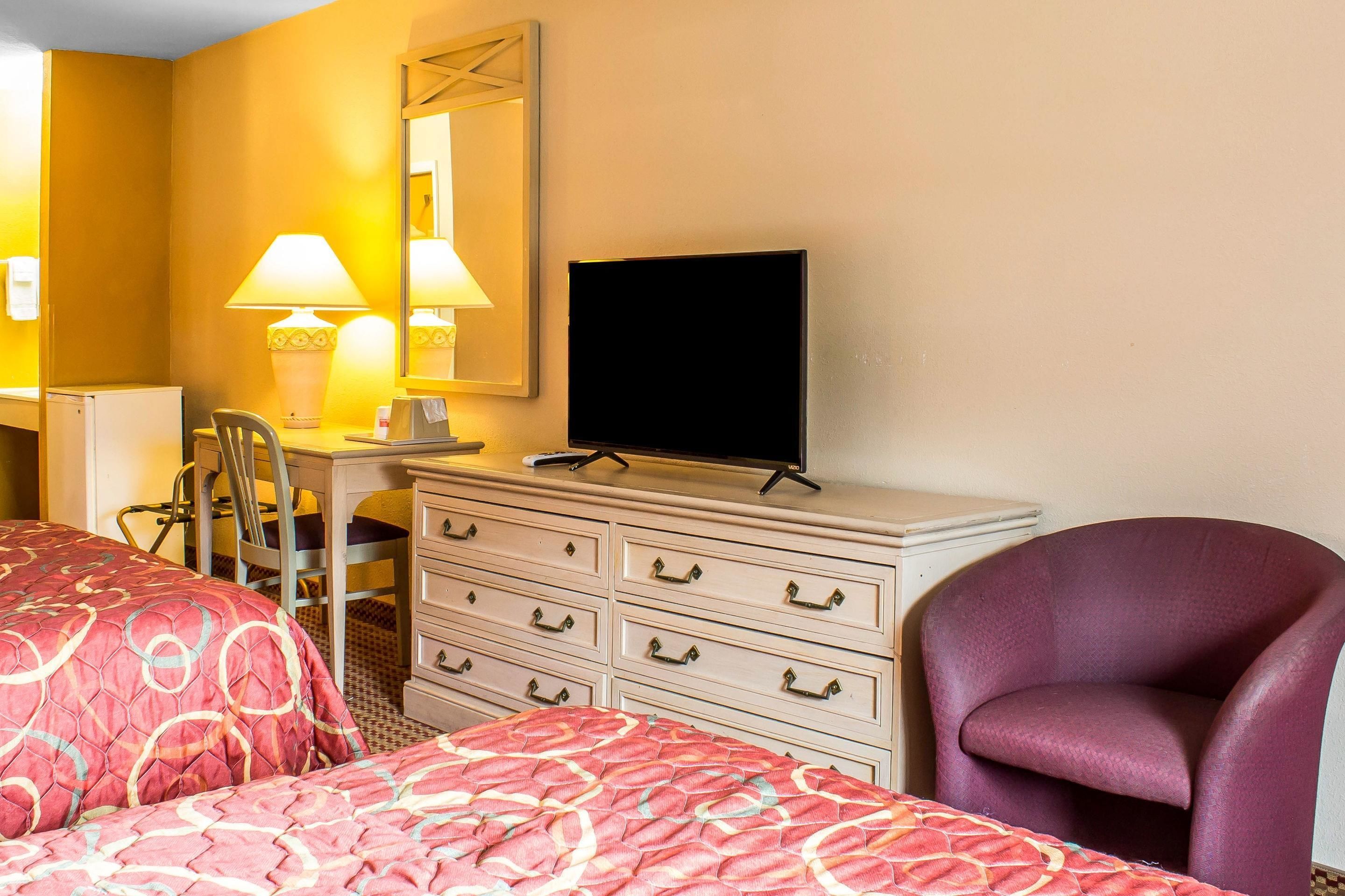 A hotel room with two beds, a desk with a chair, a dresser, a television, and a purple armchair.
