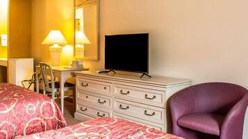A hotel room with two beds, a desk with a chair, a dresser, a television, and a purple armchair.
