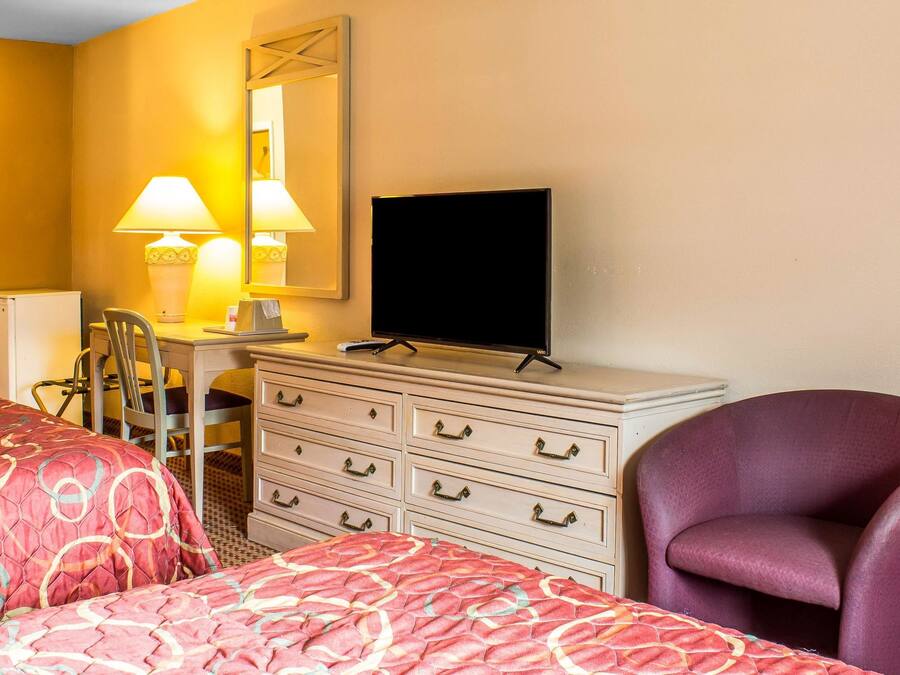 A hotel room with two beds, a desk with a chair, a dresser, a television, and a purple armchair.