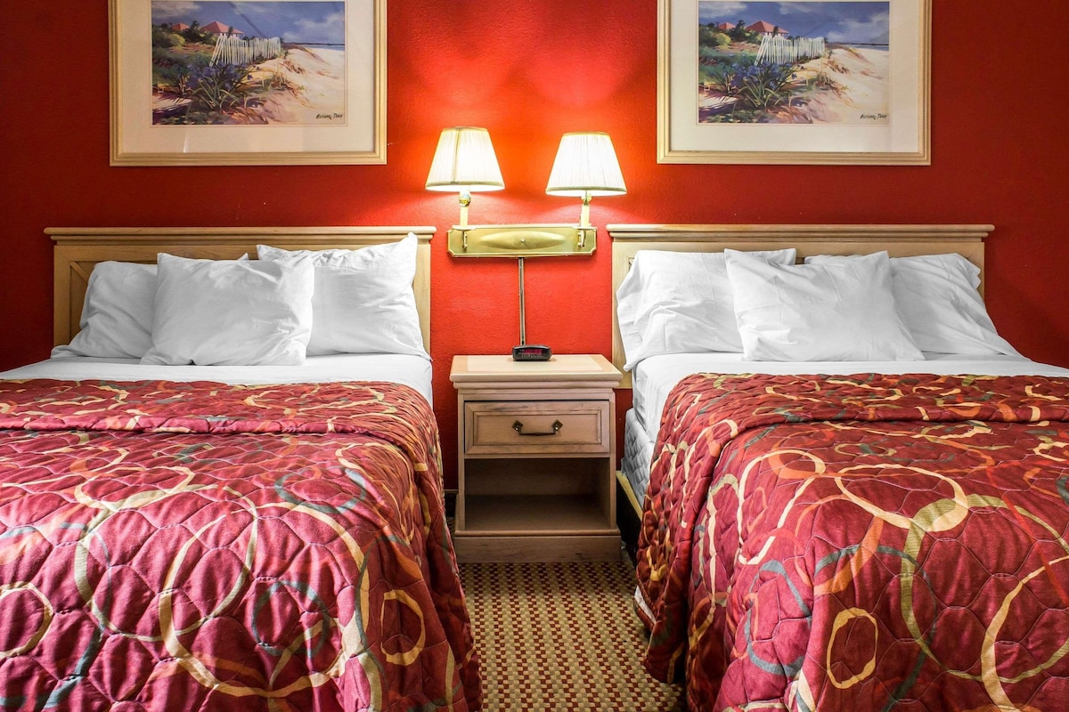 A hotel room with two beds, each with a red patterned bedspread, a nightstand with a lamp, and two framed pictures on the wall.