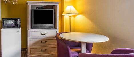 Une chambre d’hôtel compacte comprenant un coin cuisine, une télévision, une petite table, des chaises violettes et une lampe.