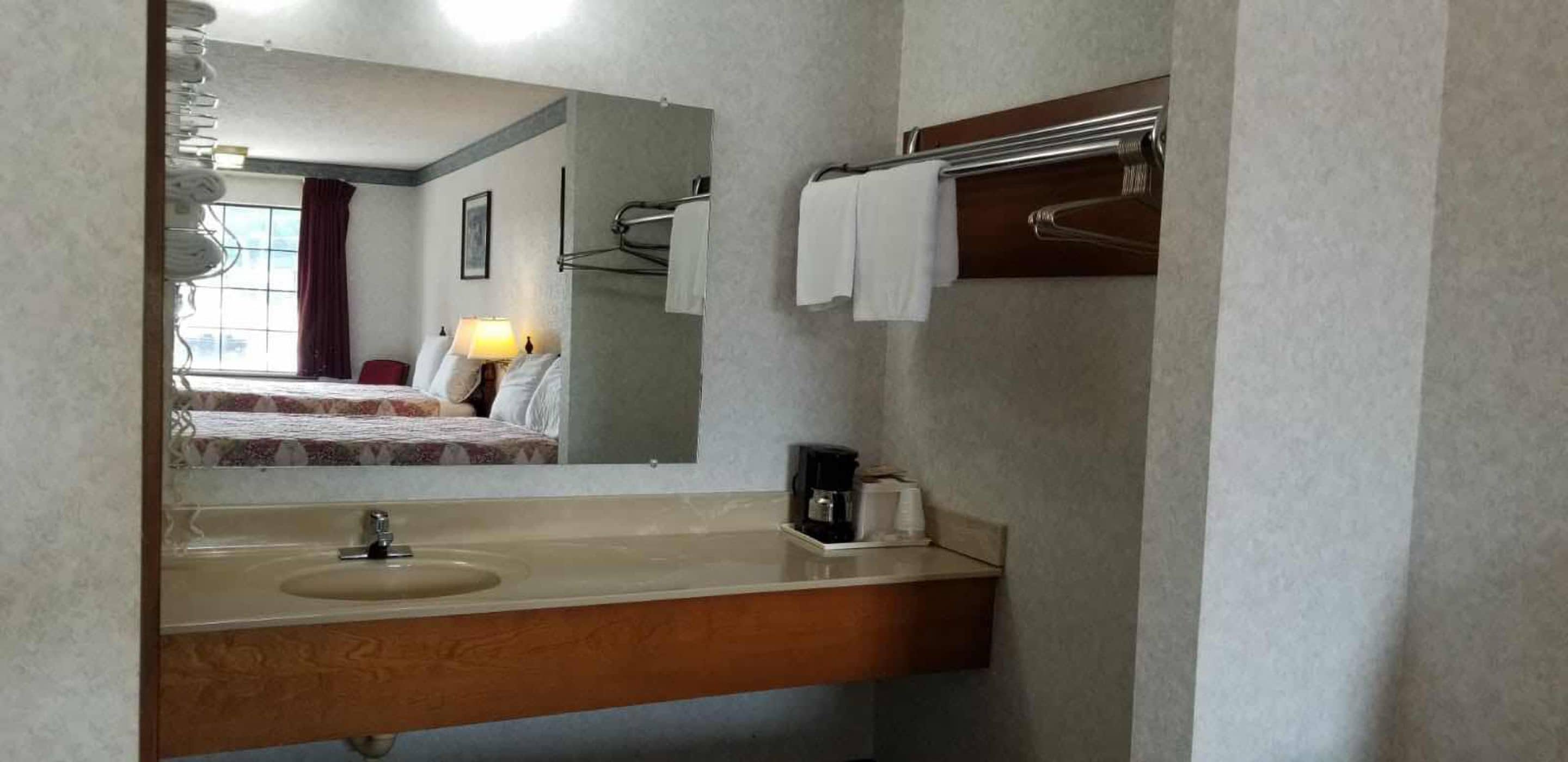Een hotelbadkamer met een wastafel, spiegel, handdoekrek en een bed zichtbaar door het raam.