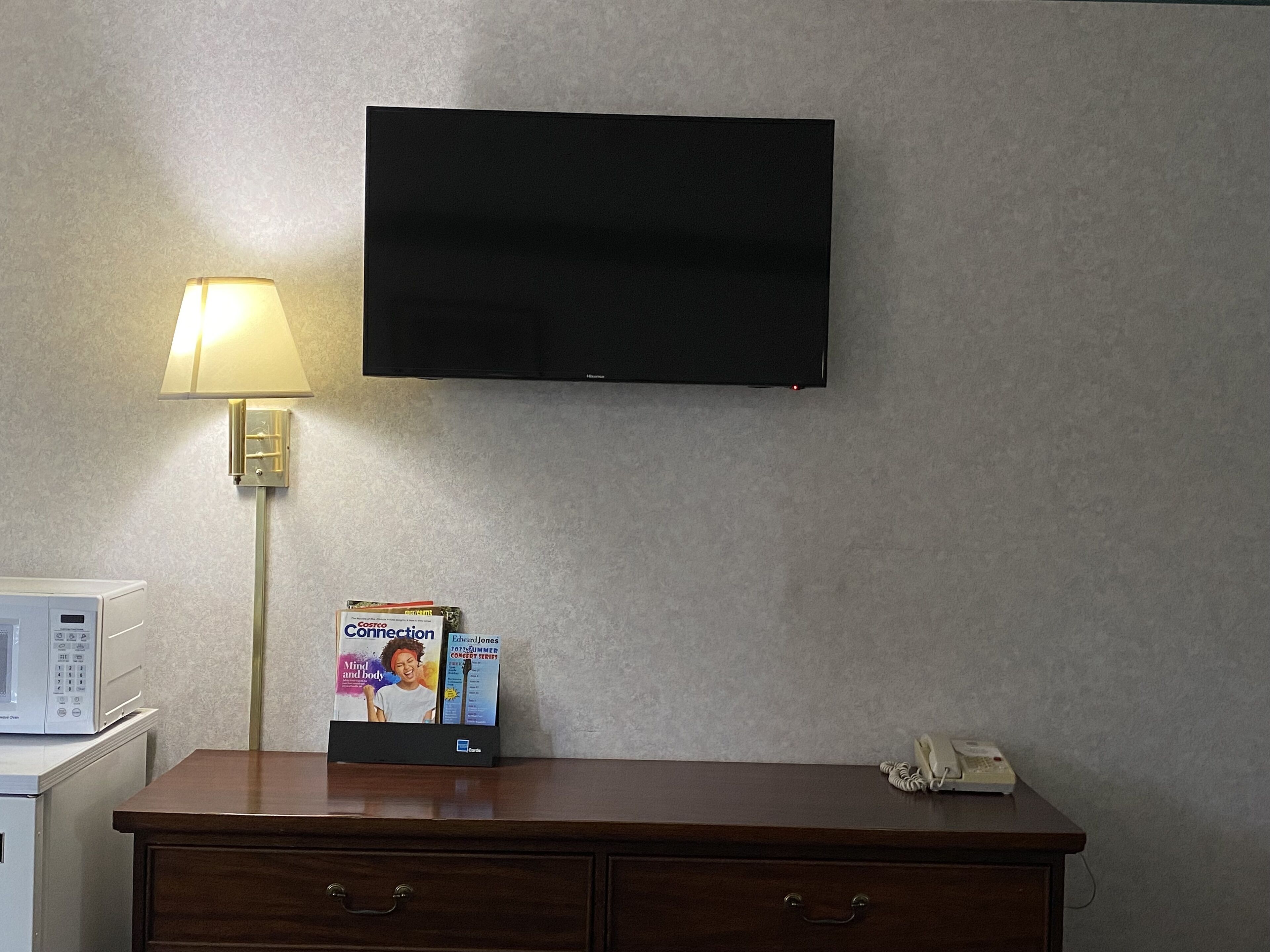 Habitación de hotel con televisión de pantalla plana en la pared, lámpara de noche, microondas, teléfono y un mueble con revistas.