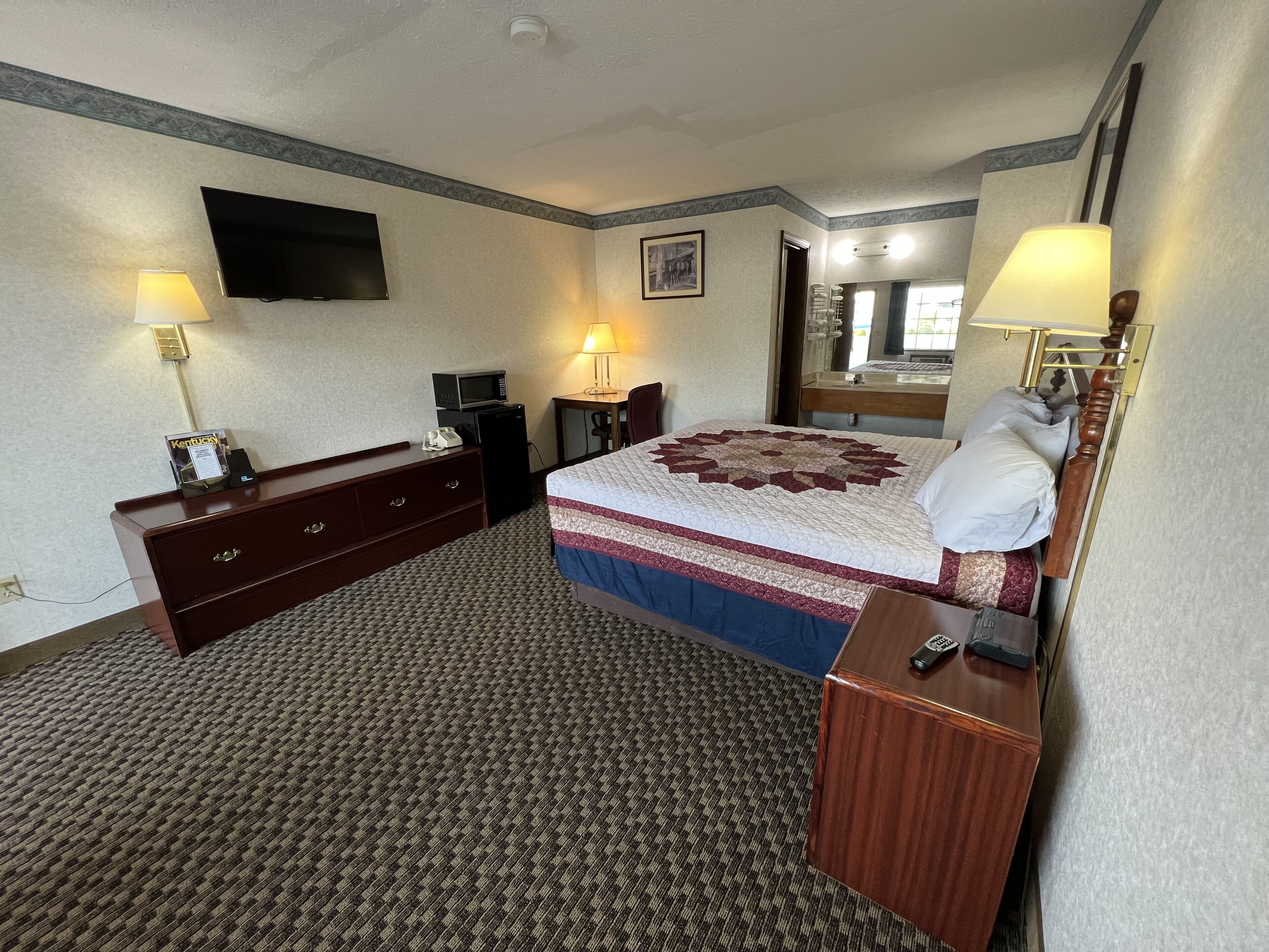 Una habitación de hotel con una cama, un escritorio, una silla, un televisor, un microondas y una mesita de noche con lámpara.