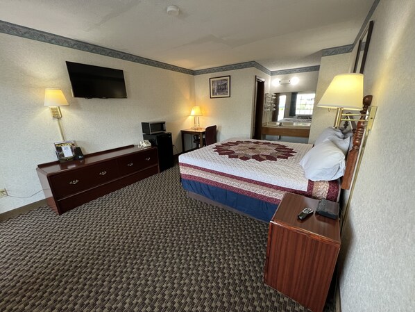 A hotel room with a bed, a desk, a chair, a television, a microwave, and a bedside table with a lamp.