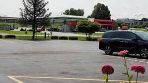 Um estacionamento com um posto de gasolina ao fundo, uma árvore e um carro estacionado em primeiro plano.