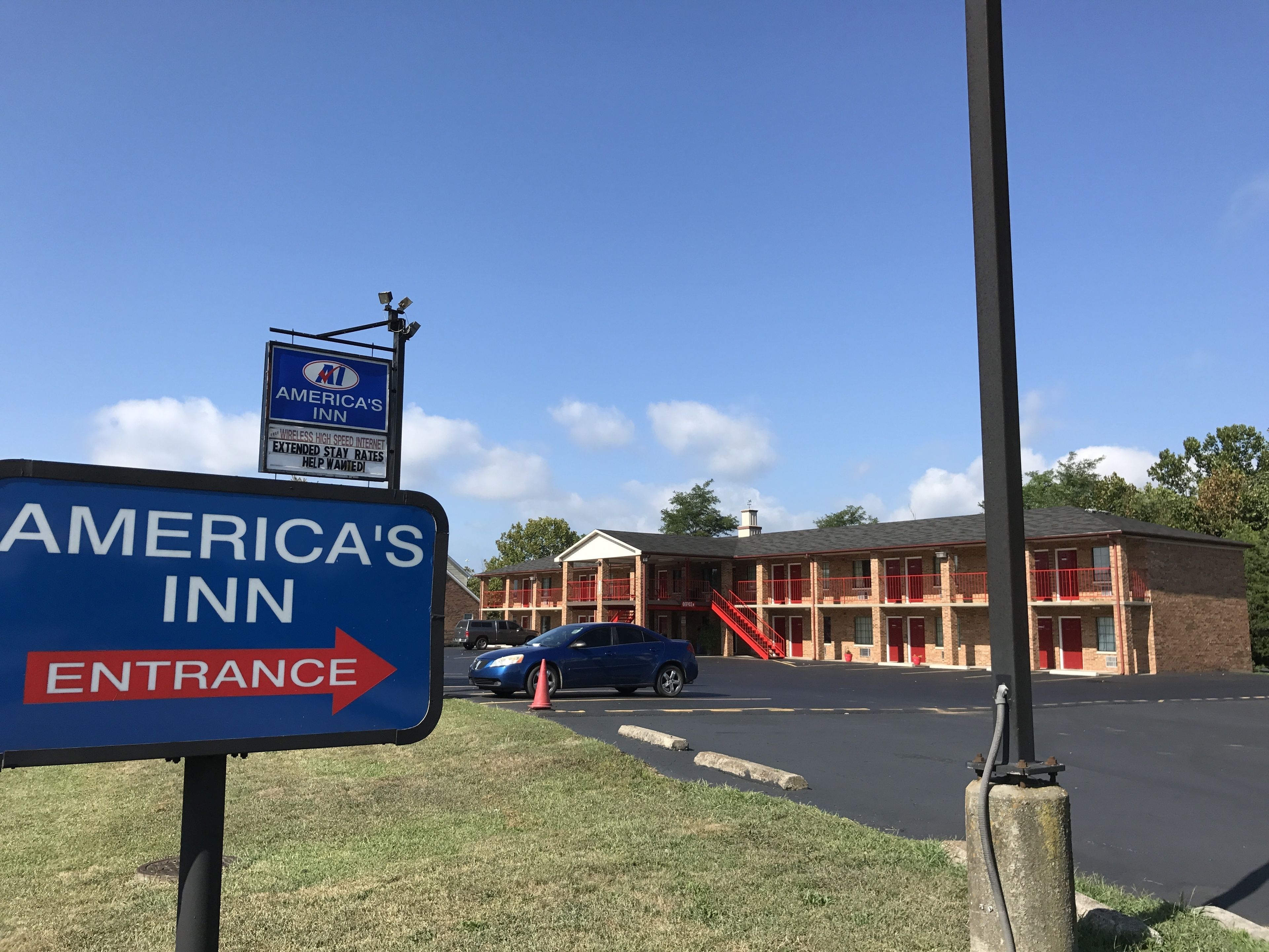 Señal de America's Inn con flecha indicando la entrada, estacionamiento y edificio de techo rojo y paredes de ladrillo.