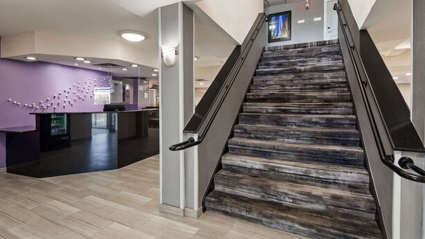 A modern hotel lobby with a reception desk, a staircase, and a purple wall with a unique design. - Best Western Paramus Hotel & Suites (Paramus)