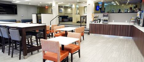 A modern hotel lobby with a reception desk, seating area with tables and chairs, and a large flat-screen TV mounted on the wall.
