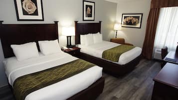 A hotel room with two beds, a desk, and a chair. There are framed pictures on the walls and a window with curtains.