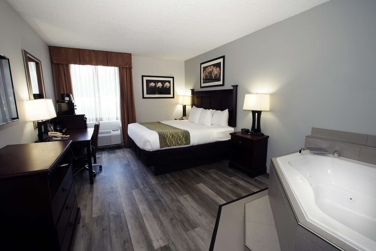 A hotel room with a bed, desk, chair, lamps, and a bathtub.