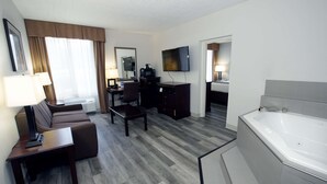 A hotel room with a sofa, a desk, a TV, a coffee maker, a desk lamp, a bathroom with a bathtub, and a large window with curtains. - Best Western Paramus Hotel & Suites (Paramus)