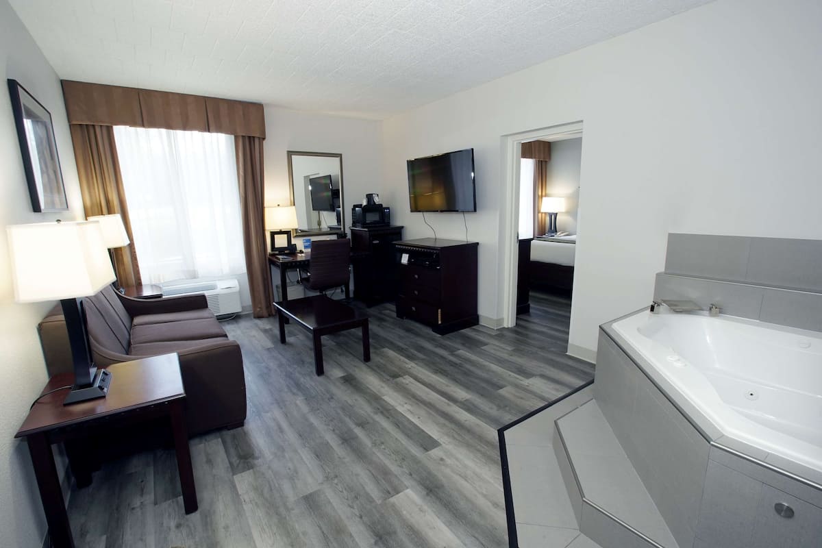 A hotel room with a sofa, a desk, a TV, a coffee maker, a desk lamp, a bathroom with a bathtub, and a large window with curtains.