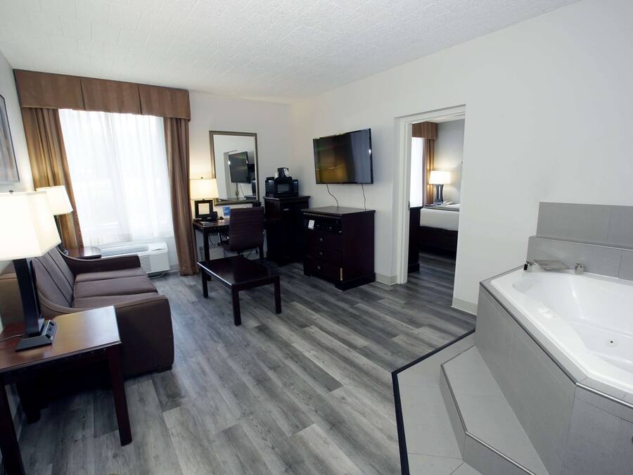 A hotel room with a sofa, a desk, a TV, a coffee maker, a desk lamp, a bathroom with a bathtub, and a large window with curtains.