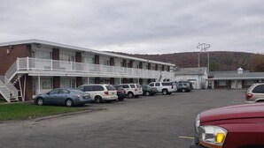 Un motel con un edificio de dos pisos, un estacionamiento con varios coches y una colina al fondo.