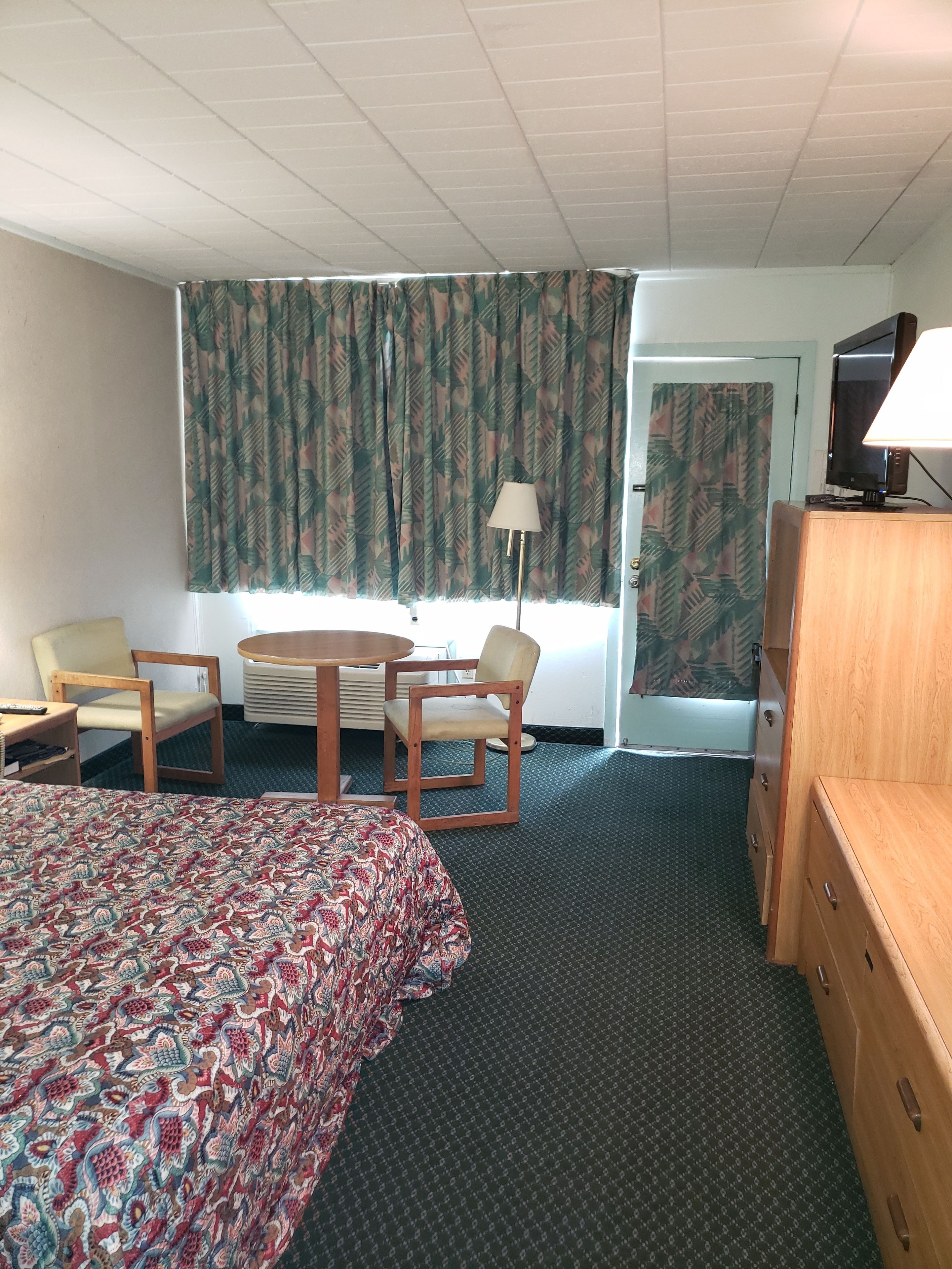 A hotel room with a bed, two chairs, a small table, a lamp, a TV, and a dresser.