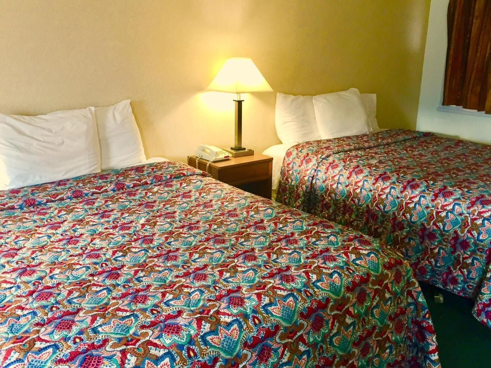 A hotel room with two beds, each with a colorful patterned bedspread. There is a bedside table with a lamp and a phone.