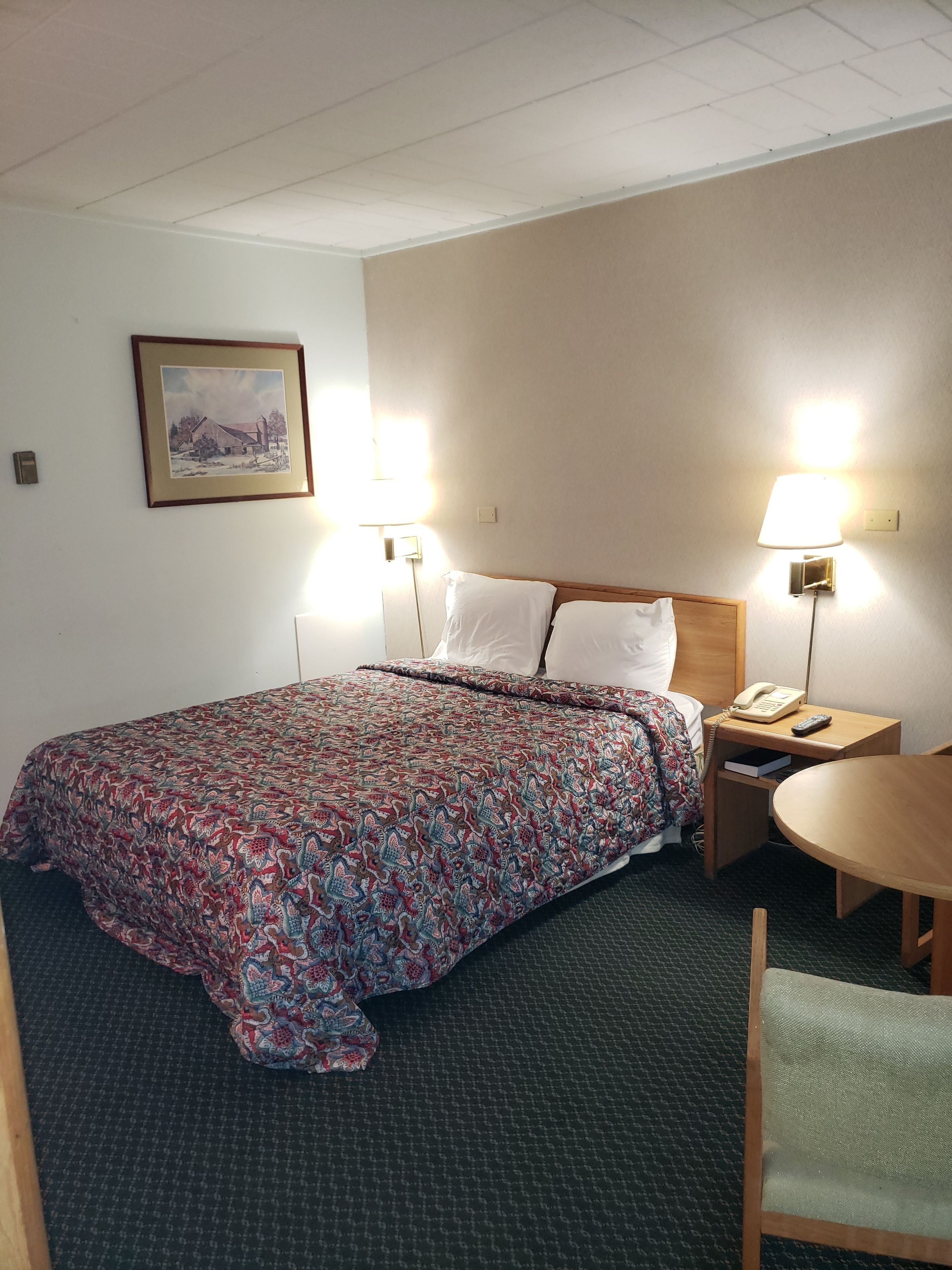 Een hotelkamer met een bed, twee bedlampen, een klein tafeltje, een stoel en een ingelijst beeld aan de muur.