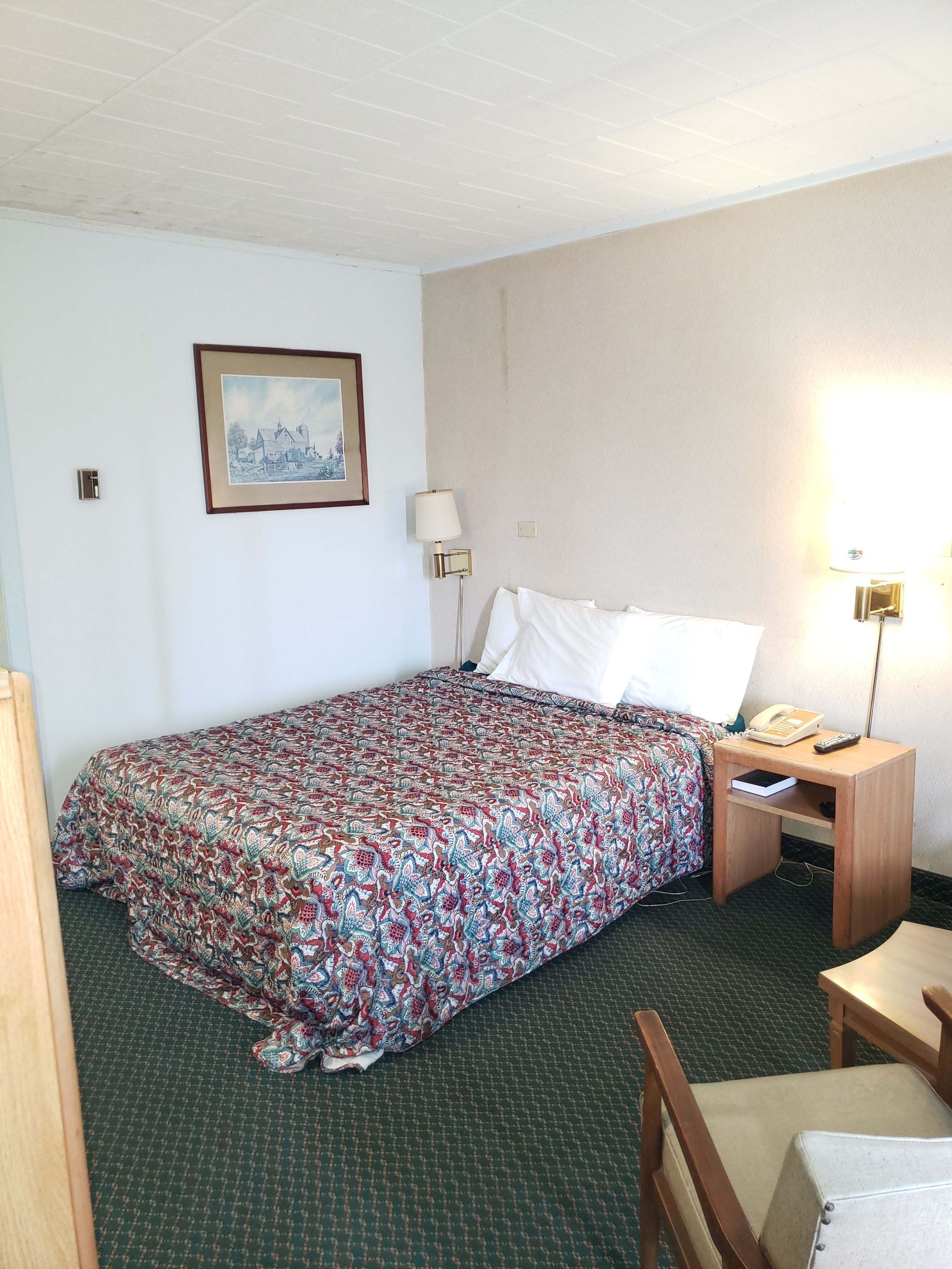 A hotel room with a bed, a bedside table with a lamp, a chair, and a framed picture on the wall.