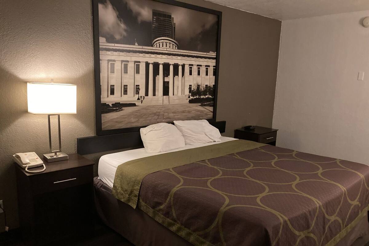 Een hotelkamer met een bed, een nachtkastje met een lamp, een telefoon en een lijst met een foto van een gebouw.