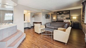A hotel room with a wooden floor, a sofa, a coffee table, a bed, a nightstand, a lamp, a painting, a mirror, a bathroom with a sink and a shower area. - Super 8 by Wyndham Millbury/Toledo (Millbury)