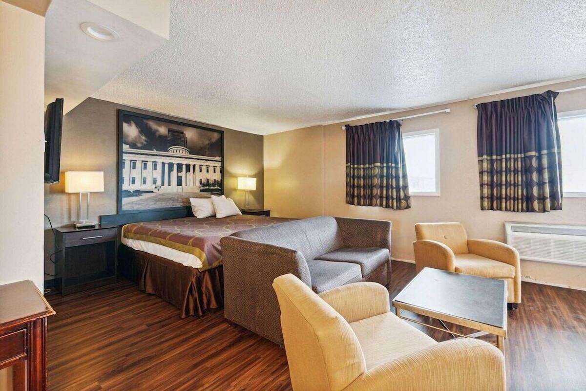 Hotelkamer met een bed, een bank, een stoel, een bijzettafeltje, een lamp, een schilderij en een raam met gordijnen.