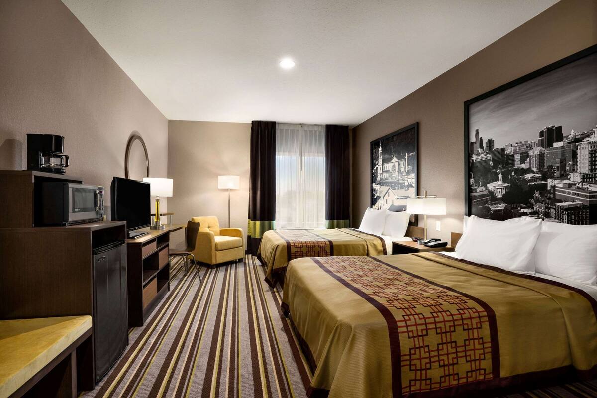 Habitación de hotel con dos camas, un escritorio con televisor, refrigerador, una silla y un cuadro de paisaje urbano en la pared.