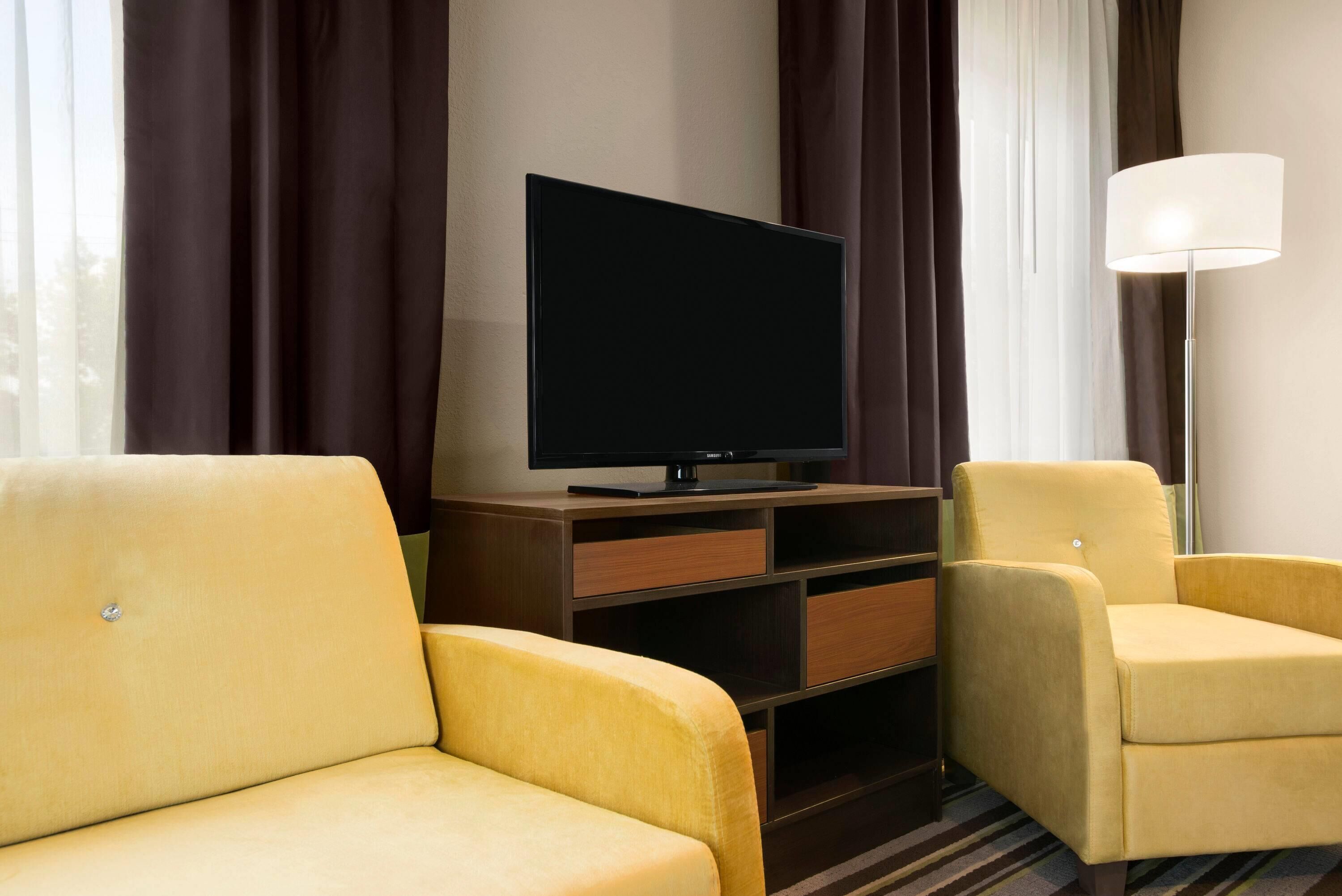 A hotel room with a flat-screen TV on a wooden stand, two yellow armchairs, a floor lamp, and a window with curtains.