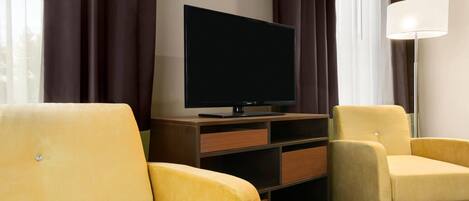 A hotel room with a flat-screen TV on a wooden stand, two yellow armchairs, a floor lamp, and a window with curtains.