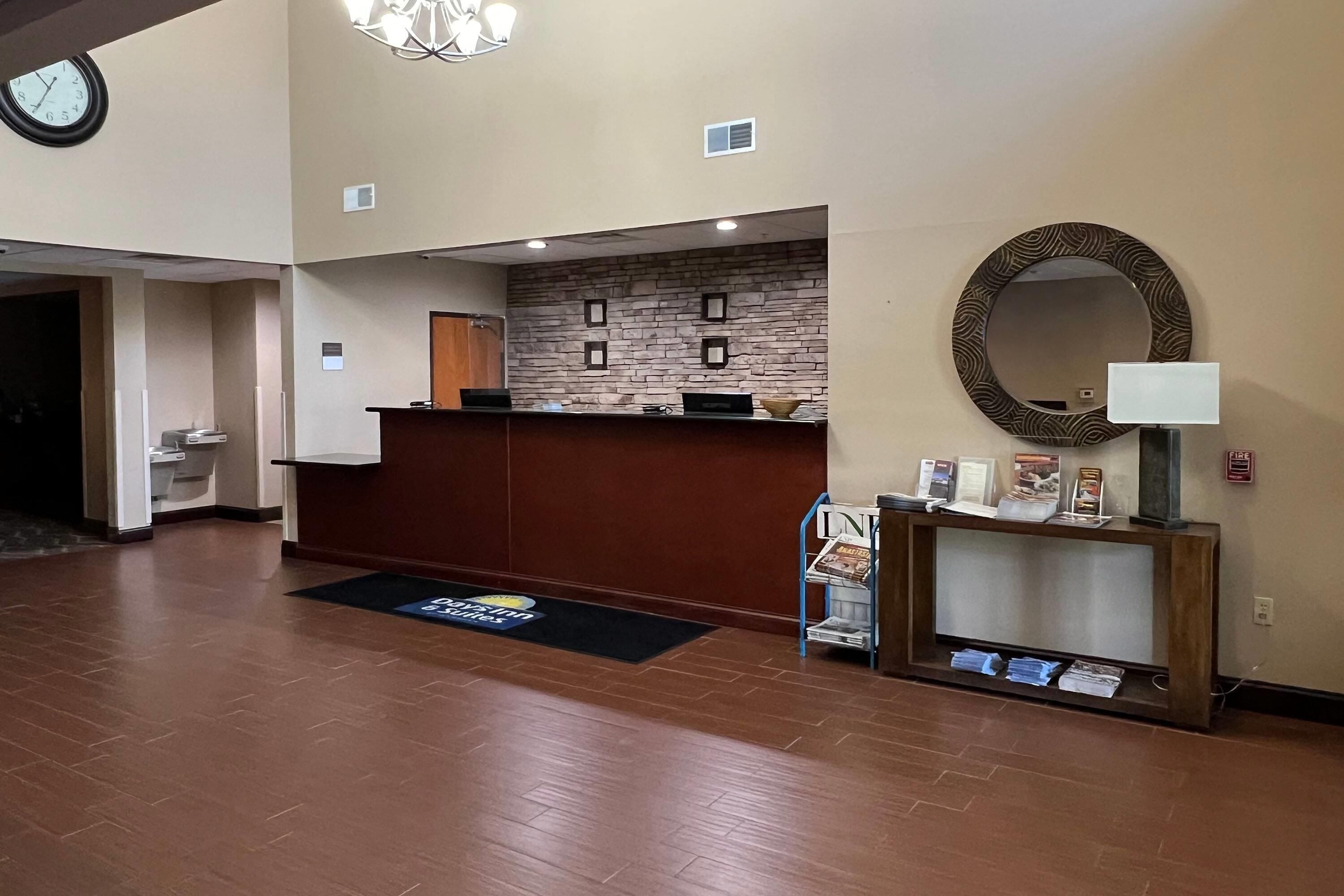 Un hall d’hôtel avec un comptoir d’accueil en bois, une horloge, un mur en pierre, un miroir rond et une petite table sur laquelle se trouvent une lampe et des brochures.