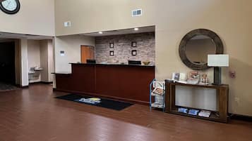Un hall d’hôtel avec un comptoir d’accueil en bois, une horloge, un mur en pierre, un miroir rond et une petite table sur laquelle se trouvent une lampe et des brochures.