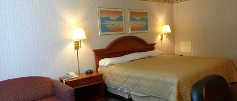 A hotel room with a bed, two bedside lamps, a telephone, a small table, a chair, and two wall-mounted paintings.