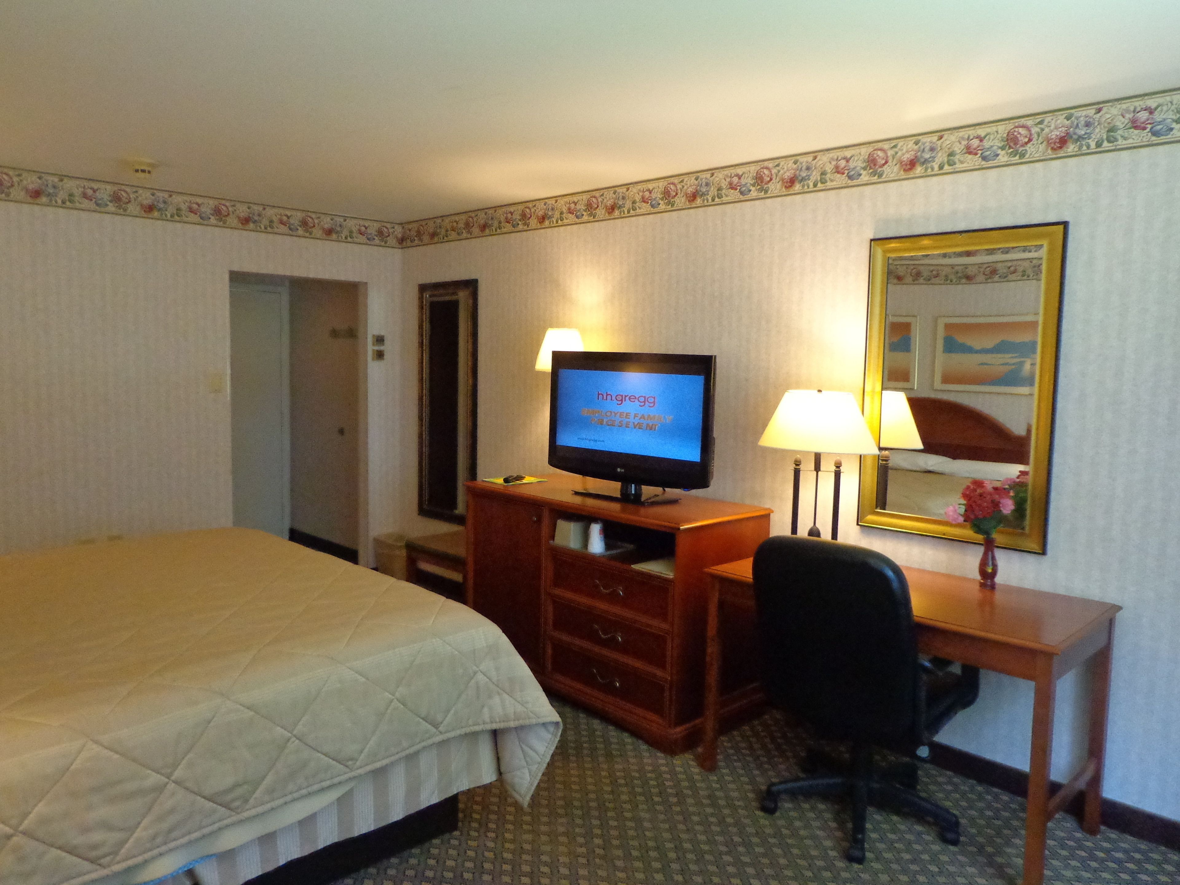 Een hotelkamer met een bed, een bureau met een computer, een stoel, een lamp en een televisie waarop 'hhgregg' staat.