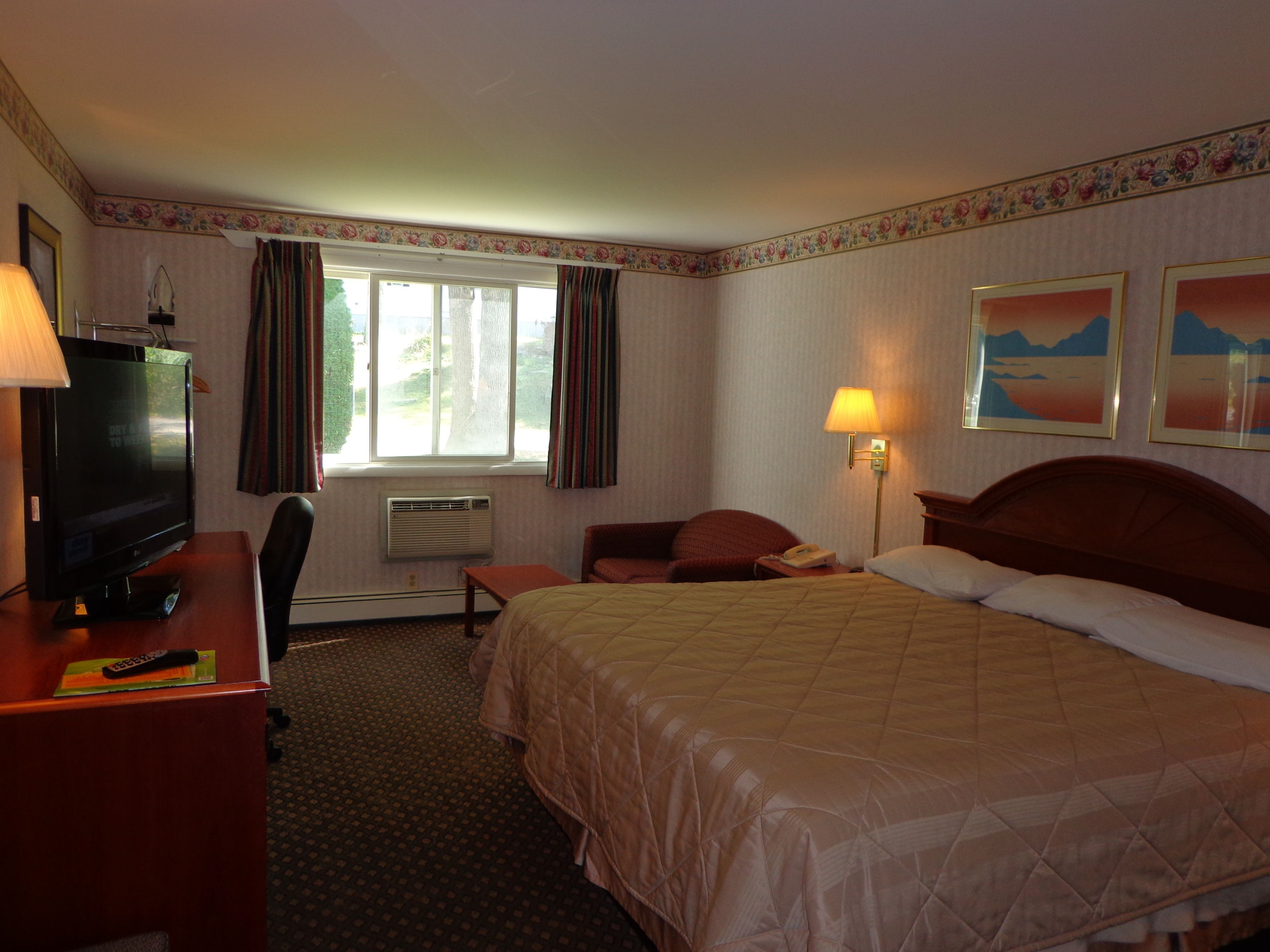 Habitación de hotel con cama, escritorio con televisor, silla, lámpara de escritorio, teléfono y ventana con cortinas.