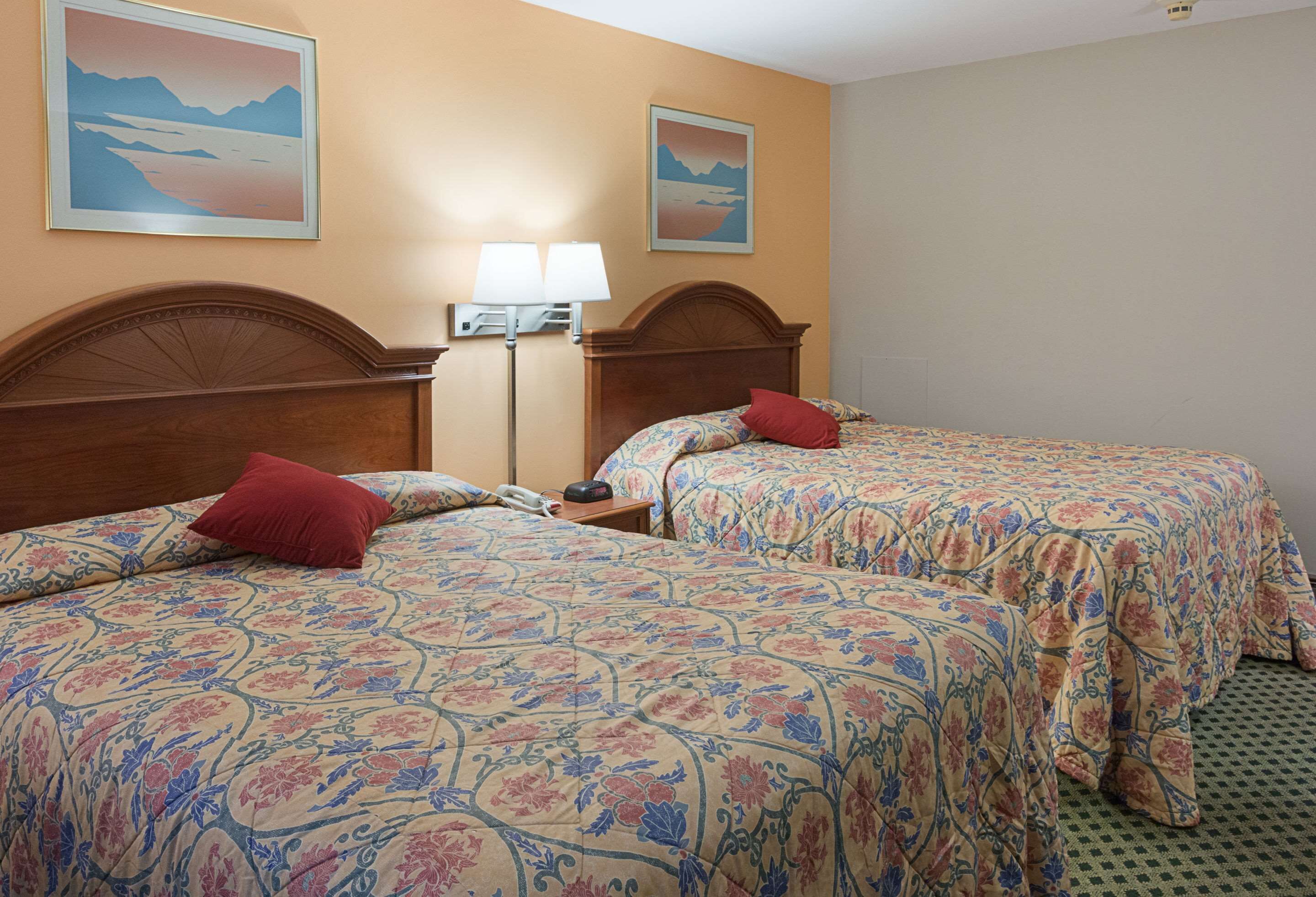 A hotel room with two beds, each with floral-patterned bedding and red pillows. There is a nightstand with a lamp and a telephone. The room has beige walls and two framed pictures on the wall.