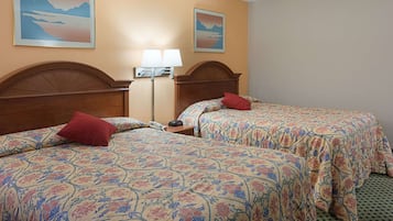 A hotel room with two beds, each with floral-patterned bedding and red pillows. There is a nightstand with a lamp and a telephone. The room has beige walls and two framed pictures on the wall.