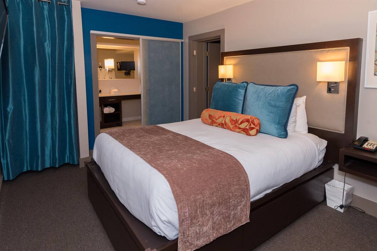 A hotel room with a bed, a nightstand, a lamp, a bathroom with a mirror, and a blue wall.
