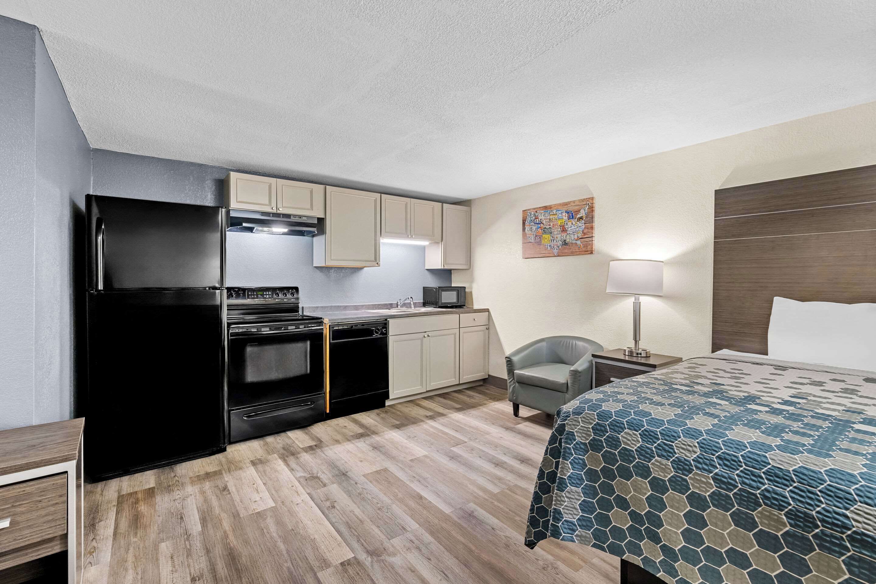 A compact hotel room with a kitchenette, a bed with a patterned bedspread, a chair, a nightstand with a lamp, and a wall art piece.