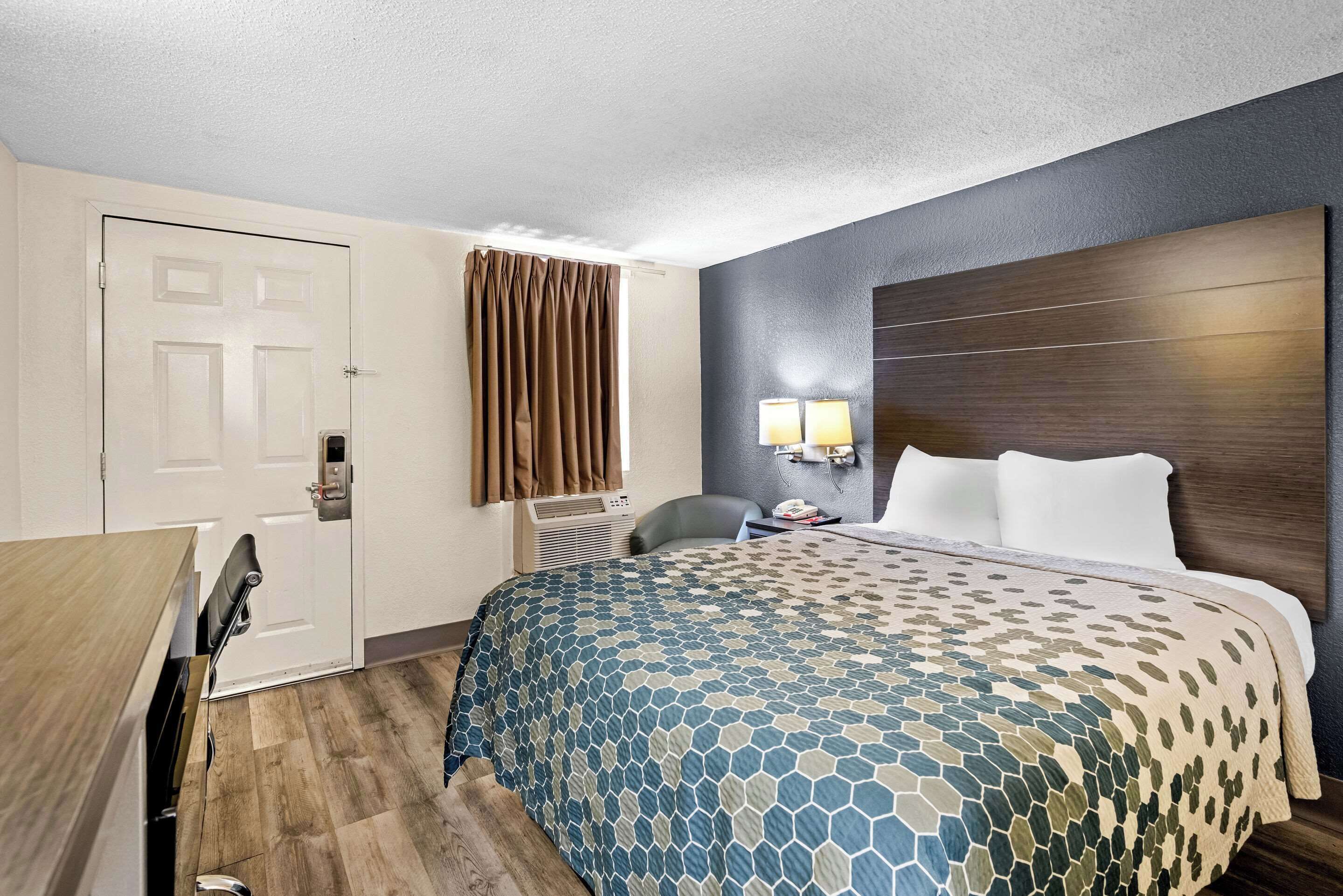 A hotel room with a bed, a desk, a chair, a window with curtains, and a wall-mounted lamp.