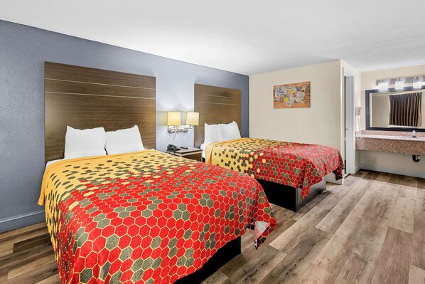 A hotel room with two beds, each with a patterned bedspread, a nightstand with a lamp, a wall-mounted artwork, and a bathroom with a mirror and towel rack.